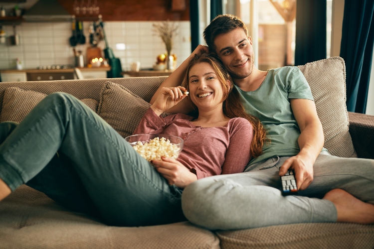 Momak i devojka sede na kauču, jedu kokice i gledaju tv
